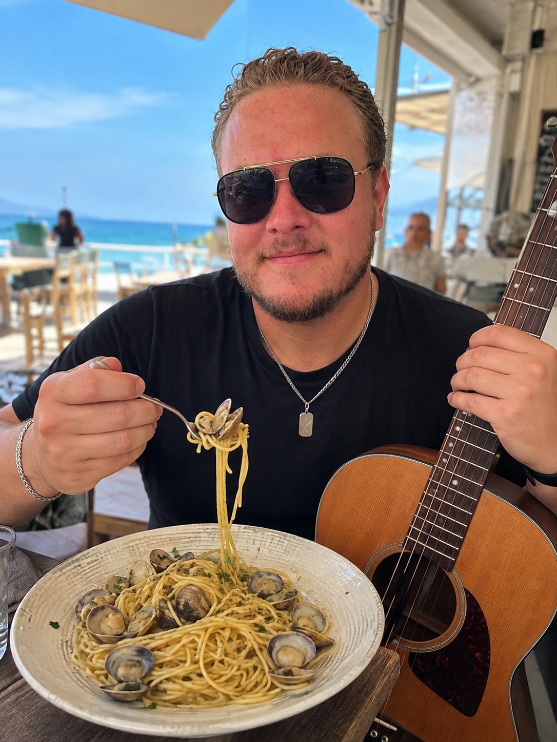 Arthur en terrasse au bord de la mer, une fourchette de spaghetti aux vongole dans une main, sa guitare dans l'autre